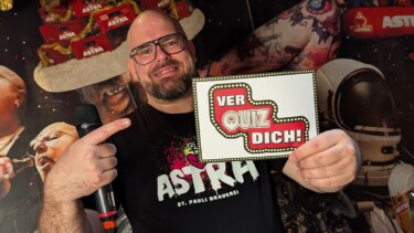 Person hält Mikrofon und Schild mit Aufschrift 'VER QUIZ DICH!' vor einem Hintergrund mit Astra-Logos und Weihnachtsdekoration.