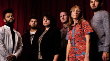 Gruppe von sechs Personen vor rotem Vorhang, eine trägt ein rotes Hemd mit blauem Lippenmuster, andere in dunkler und heller Kleidung.