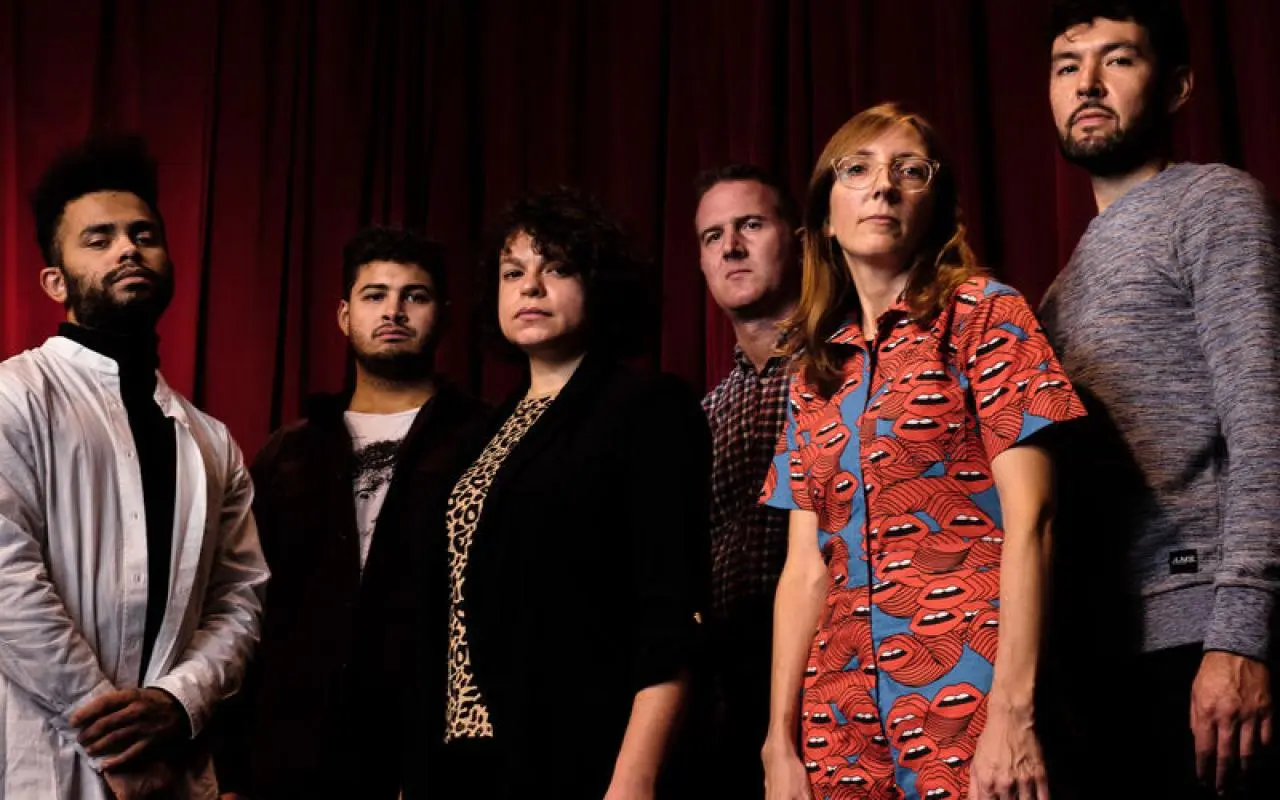 Gruppe von sechs Personen vor rotem Vorhang, eine trägt ein rotes Hemd mit blauem Lippenmuster, andere in dunkler und heller Kleidung.