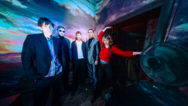 Fünf Personen in einem Raum mit farbiger Wandprojektion, eine Person trägt roten Pullover und streckt einen Arm aus.