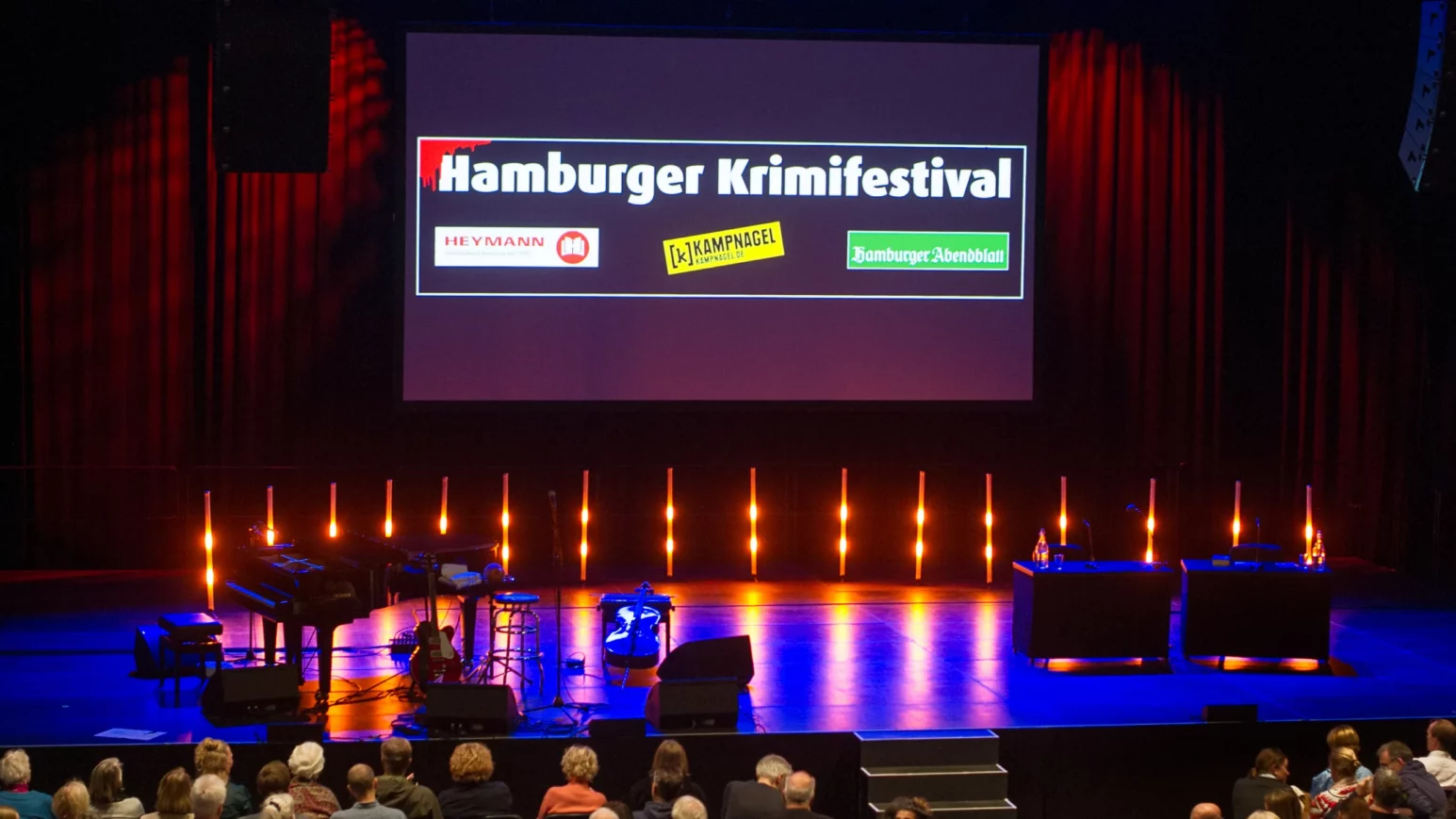 Bühne mit Instrumenten und Mikrofonen vor Leinwand mit Schrift 'Hamburger Krimifestival' und Logos von Heymann, Krimispiegel, Hamburger Abendblatt.