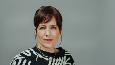 Frau mit braunen Haaren, schwarzem Pullover mit weißem Muster und goldener Halskette vor grauem Hintergrund.