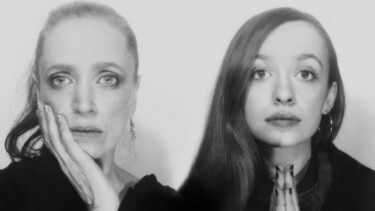 Zwei Frauen mit langen Haaren, eine mit Zopf, die andere mit offenem Haar, beide in dunkler Kleidung, Hände vor der Brust gefaltet.