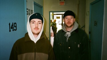 Zwei Personen in dunklen Jacken stehen in einem Flur mit blauen Türen und schwarzen Zahlen an den Wänden.