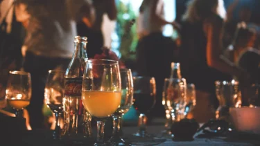 Glas mit orangefarbener Flüssigkeit und Coca-Cola-Flasche auf einem Tisch mit weiteren Gläsern und unscharfen Personen im Hintergrund.