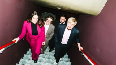 Vier Personen steigen eine Treppe mit roten Handläufen hinauf, zwei tragen Anzüge, eine Person trägt einen pinkfarbenen Anzug.