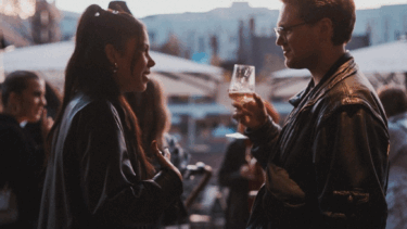 Zwei Personen in Lederjacken unterhalten sich, eine hält ein Glas mit Getränk, im Hintergrund weitere Menschen und Außenbereich mit Sonnenschirmen.