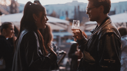 Zwei Personen in Lederjacken unterhalten sich, eine hält ein Glas mit Getränk, im Hintergrund weitere Menschen und Außenbereich mit Sonnenschirmen.