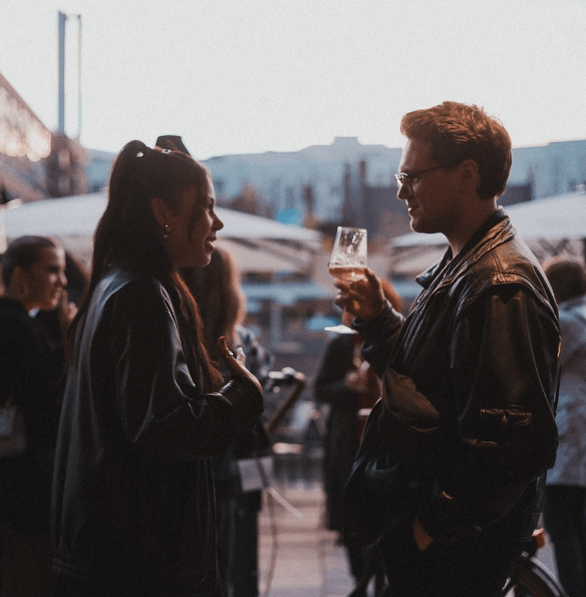 Zwei Personen in Lederjacken unterhalten sich, eine hält ein Glas mit Getränk, im Hintergrund weitere Menschen und Außenbereich mit Sonnenschirmen.