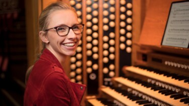 Person mit rotem Jackett sitzt an einer Orgel mit mehreren Tastaturen und Registerknöpfen, Notenblatt aufgestellt.