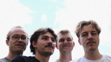 Vier Personen stehen vor hellem Himmel, tragen helle T-Shirts und blicken nach oben.