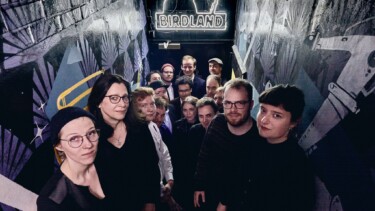 Gruppe von Personen in einem schmalen, dunklen Gang mit Wandmalereien und einem beleuchteten Schild mit der Aufschrift 'BIRDLAND'.