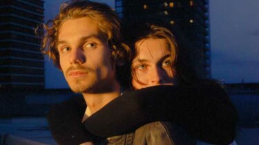 Zwei Personen umarmen sich, eine trägt eine schwarze Lederjacke und silbernen Schmuck an der Hand, im Hintergrund zwei hohe Gebäude bei Dämmerung.