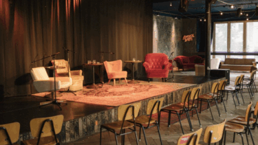 Bühne mit verschiedenen Sesseln und Teppich, davor Reihen von Holzstühlen in einem Veranstaltungsraum mit großen Fenstern.