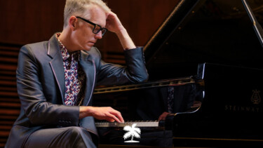 Person mit kurzem blondem Haar in dunklem Anzug sitzt am schwarzen Steinway-Flügel und berührt die Klaviatur mit einer Hand.