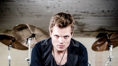 Mann mit schwarzem T-Shirt und Weste sitzt vor Schlagzeug, Hände verschränkt.