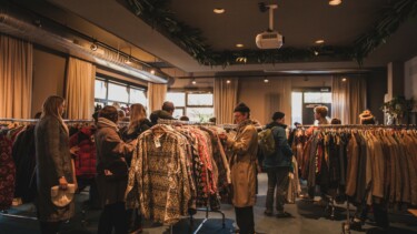 Menschen in einem Geschäft betrachten Kleidung an Kleiderständern unter warmem Licht und mit Vorhängen an den Fenstern.