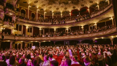 Voller Zuschauerraum in einem historischen Theater mit zwei Rängen und reich verzierten Balkonen, einige Personen halten Bewertungsplakate hoch.