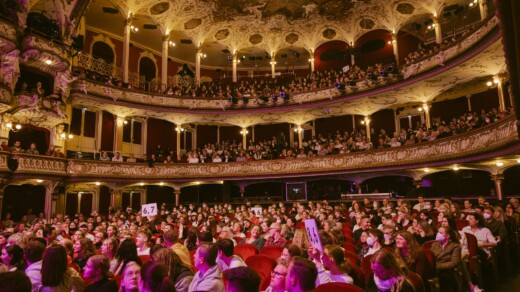 Voller Zuschauerraum in einem historischen Theater mit zwei Rängen und reich verzierten Balkonen, einige Personen halten Bewertungsplakate hoch.