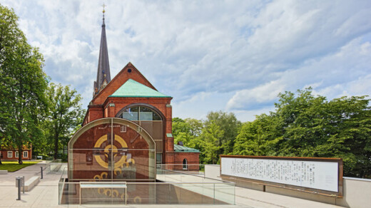 Rotes Backsteingebäude mit spitzem Turm und Glasvitrine mit goldfarbenem Zifferblatt vor grünem Baumhintergrund.
