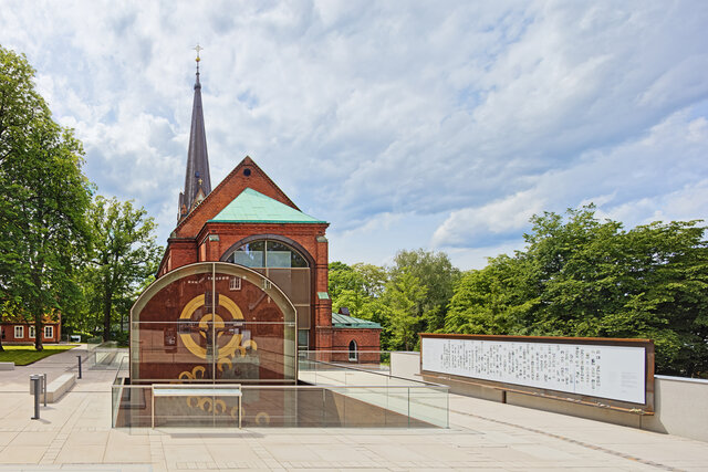 Rotes Backsteingebäude mit spitzem Turm und Glasvitrine mit goldfarbenem Zifferblatt vor grünem Baumhintergrund.