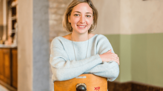 Person mit hellblondem, schulterlangem Haar trägt hellblauen Pullover und sitzt mit verschränkten Armen auf einem Holzstuhl