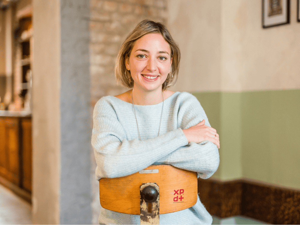 Person mit hellblondem, schulterlangem Haar trägt hellblauen Pullover und sitzt mit verschränkten Armen auf einem Holzstuhl