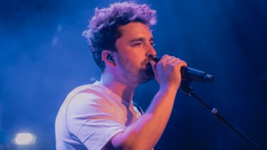 Person mit lockigem Haar und weißem T-Shirt singt in ein Mikrofon auf einer Bühne mit blauem Bühnenlicht.