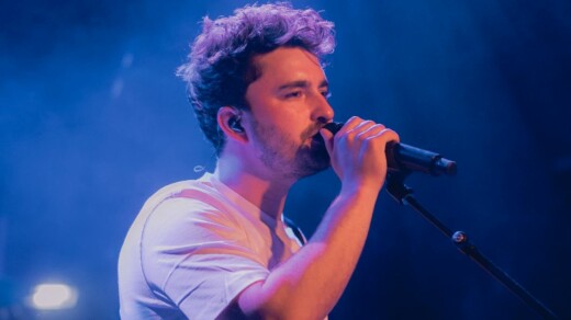 Person mit lockigem Haar und weißem T-Shirt singt in ein Mikrofon auf einer Bühne mit blauem Bühnenlicht.