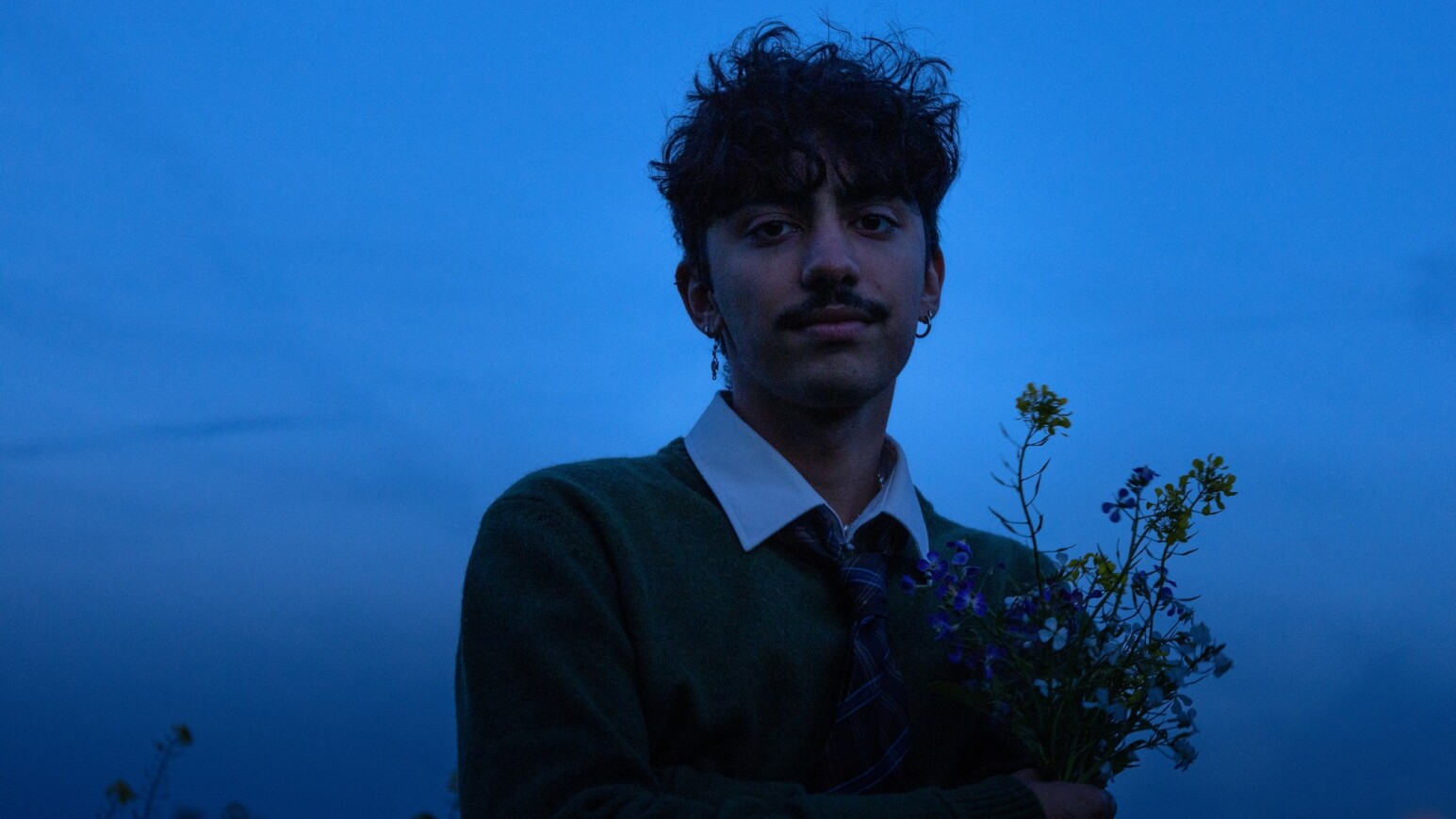 Person mit dunklem Pullover und weißem Hemd hält einen kleinen Strauß Wildblumen vor blauem Abendhimmel.
