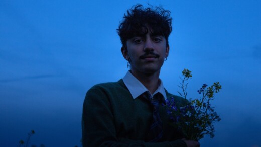Person mit dunklem Pullover und weißem Hemd hält einen kleinen Strauß Wildblumen vor blauem Abendhimmel.