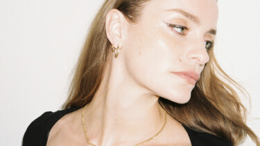 Frau mit langen blonden Haaren, trägt goldene Creolen und eine dünne goldene Halskette, schwarzes Oberteil sichtbar.