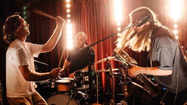 Band spielt live auf Bühne mit Schlagzeug, Saxofon und E-Gitarre vor rotem Vorhang und Bühnenbeleuchtung.