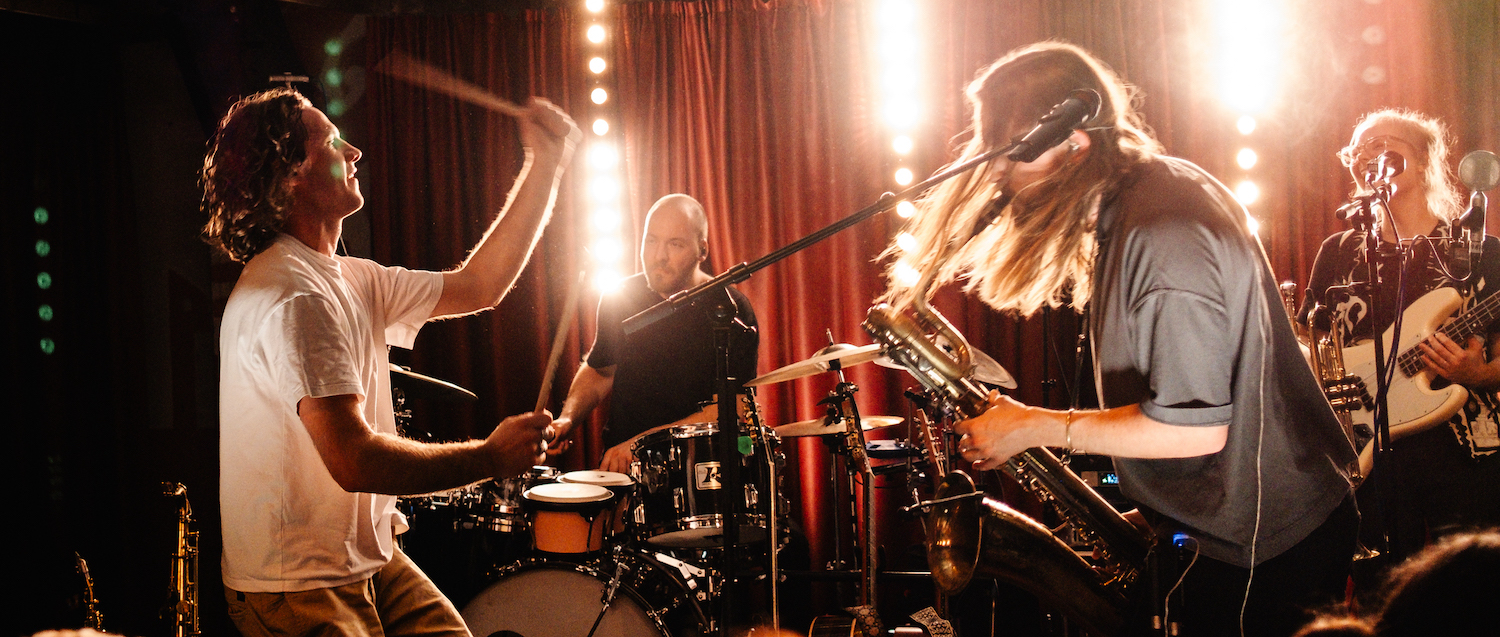 Band spielt live auf Bühne mit Schlagzeug, Saxofon und E-Gitarre vor rotem Vorhang und Bühnenbeleuchtung.