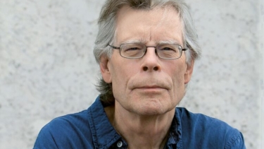 Älterer Mann mit grauen Haaren, blauem Hemd, sitzt vor einer hellen Wand.