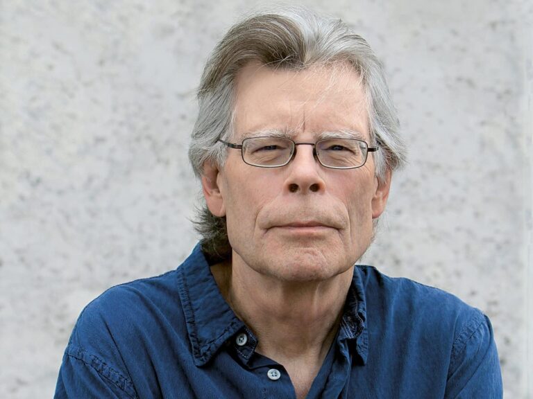 Älterer Mann mit grauen Haaren, blauem Hemd, sitzt vor einer hellen Wand.