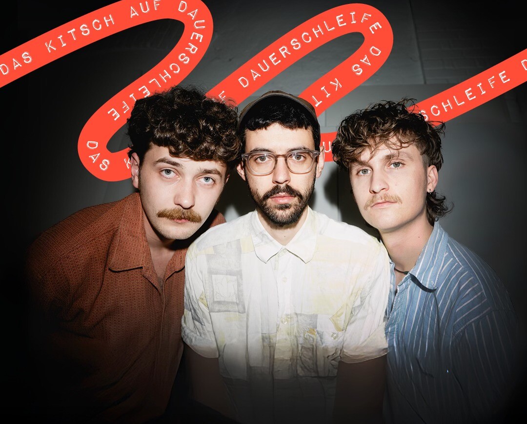 Drei Personen in Hemden vor schwarzer Wand mit rotem Schriftband, darauf steht 'DAS KITSCH AUF DAUERSCHLEIFE'.