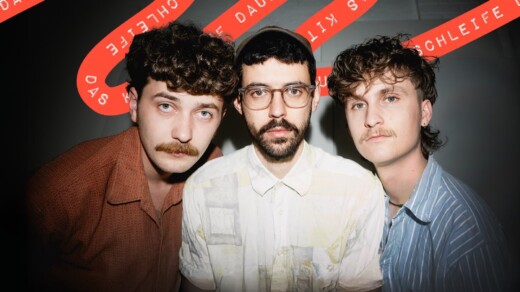 Drei Personen in Hemden vor schwarzer Wand mit rotem Schriftband, darauf steht 'DAS KITSCH AUF DAUERSCHLEIFE'.