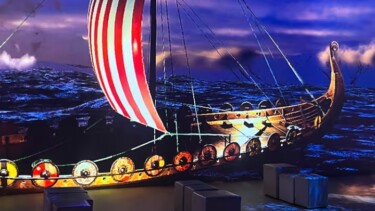 Wikingerschiff mit rot-weiß gestreiftem Segel und Rundschilden am Rumpf, dargestellt auf einer Leinwand in einem dunklen Raum mit Sitzbänken.