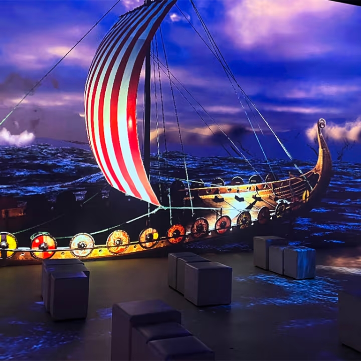 Wikingerschiff mit rot-weiß gestreiftem Segel und Rundschilden am Rumpf, dargestellt auf einer Leinwand in einem dunklen Raum mit Sitzbänken.