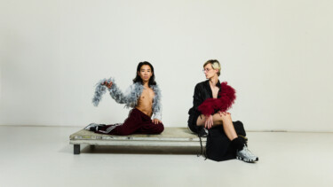 Die Hamburger Künstler:innen Lisa Rykena und Carolin Jüngst sitzen auf einer Bank vor einem weißen Hintergrund